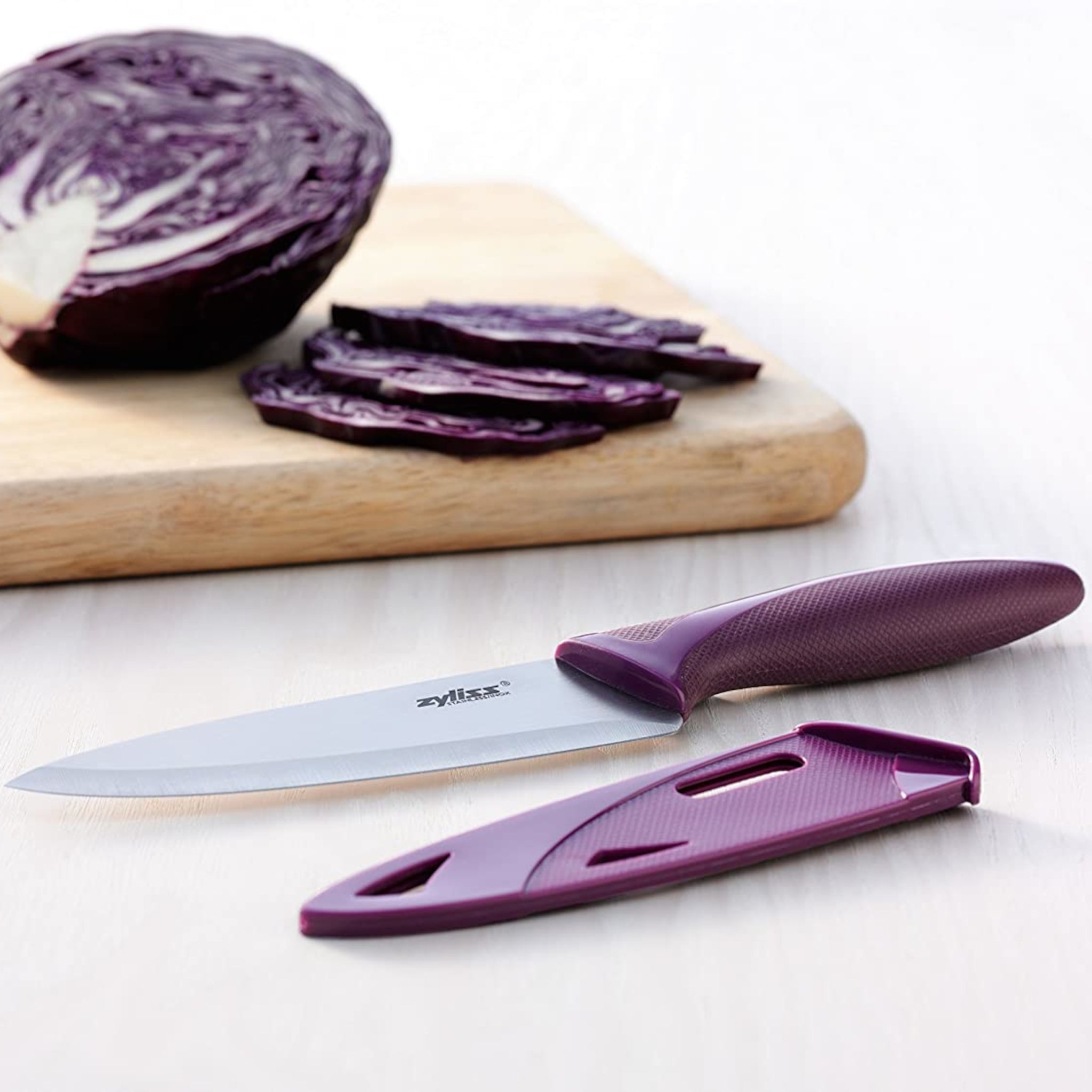 A chef’s knife with a purple handle and matching sheath lies on a white surface, next to sliced purple cabbage on a wooden cutting board.
