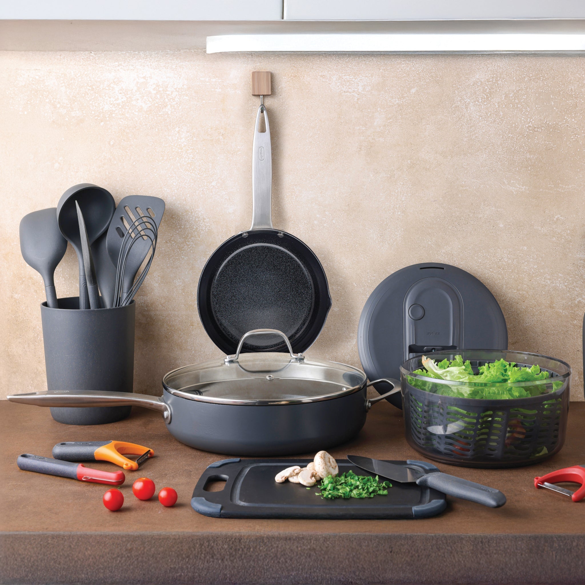 A kitchen counter with cooking utensils, pans, a salad spinner with lettuce, a cutting board with mushrooms and herbs, a knife, tomatoes, and kitchen tools neatly arranged.