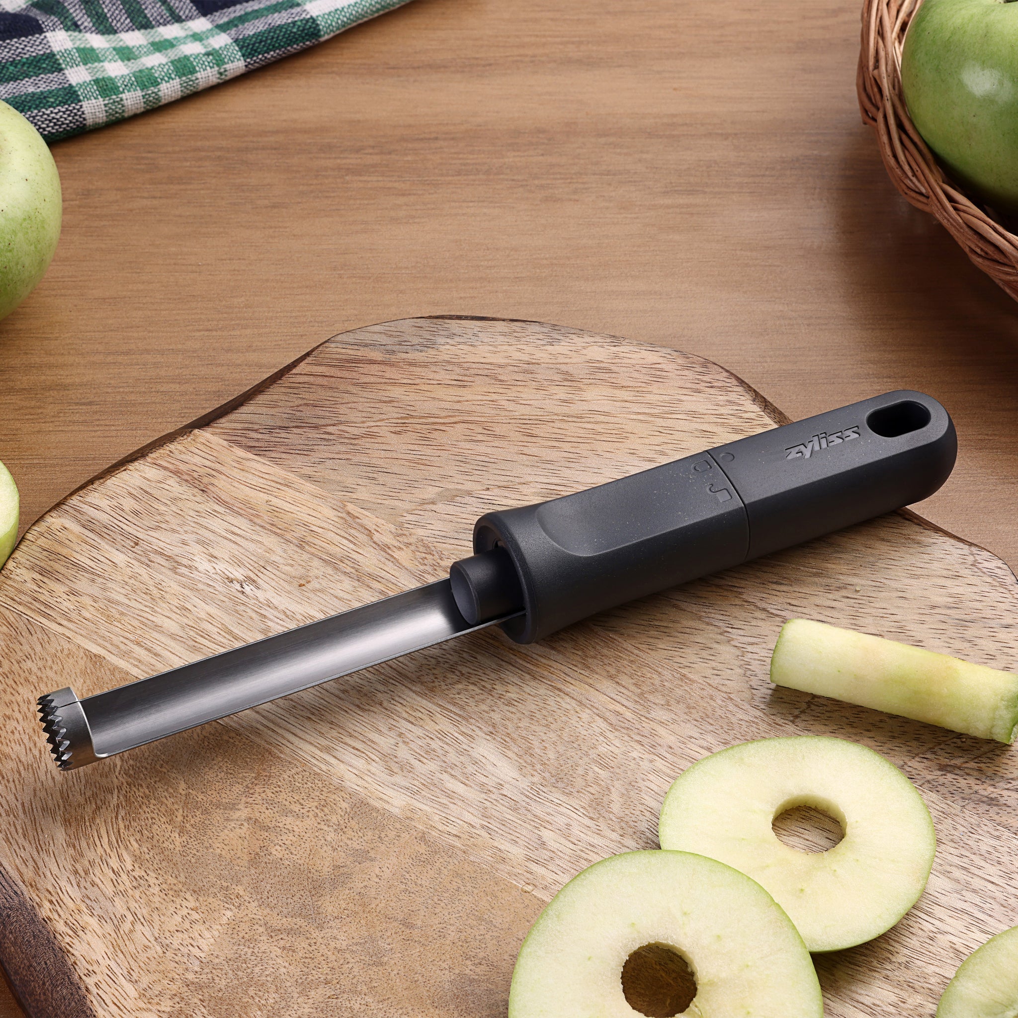 A metal apple corer with a black handle rests on a wooden cutting board. Sliced green apples and apple cores are around it, and theres a basket with more green apples. A checkered cloth is partially visible.