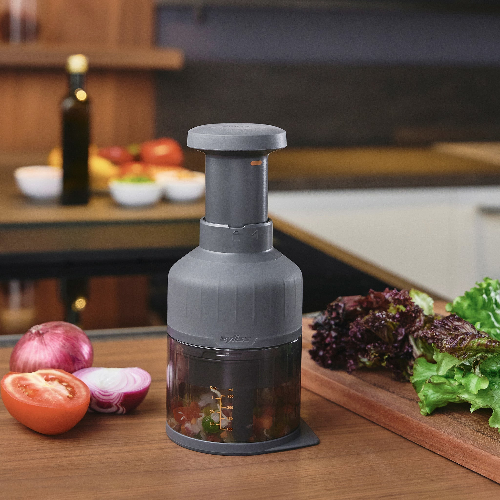A manual food chopper filled with chopped vegetables sits on a kitchen counter, with whole vegetables and a cutting board in the background.