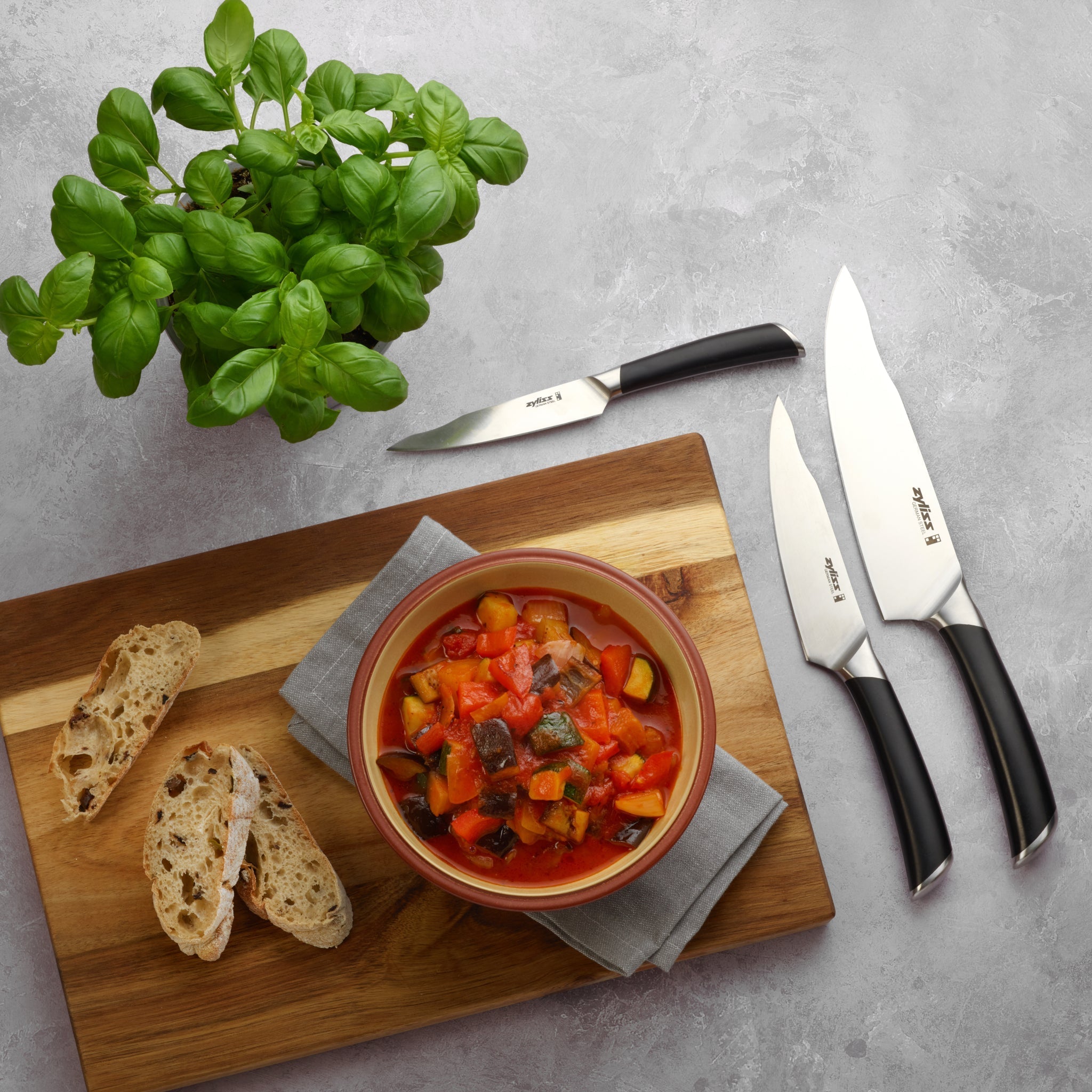A bowl of vegetable stew sits on a wooden board with two slices of bread, three kitchen knives, and a potted basil plant on a gray surface.