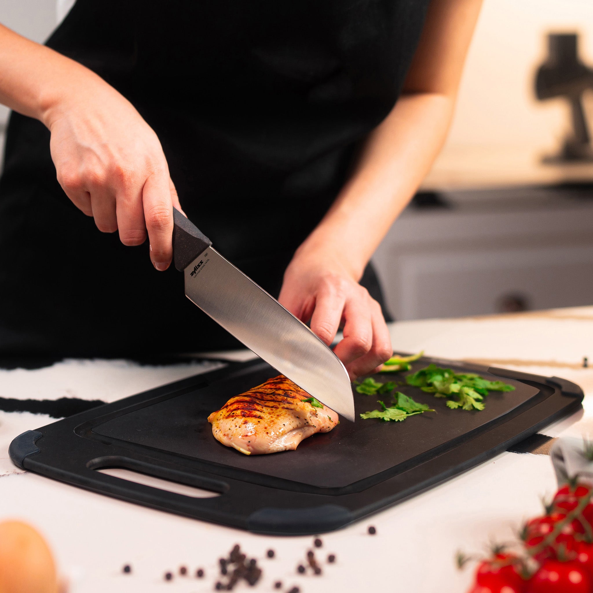 A person slices grilled chicken on a black cutting board, garnished with cilantro. Nearby, there are black peppercorns, a tomato, and an egg.