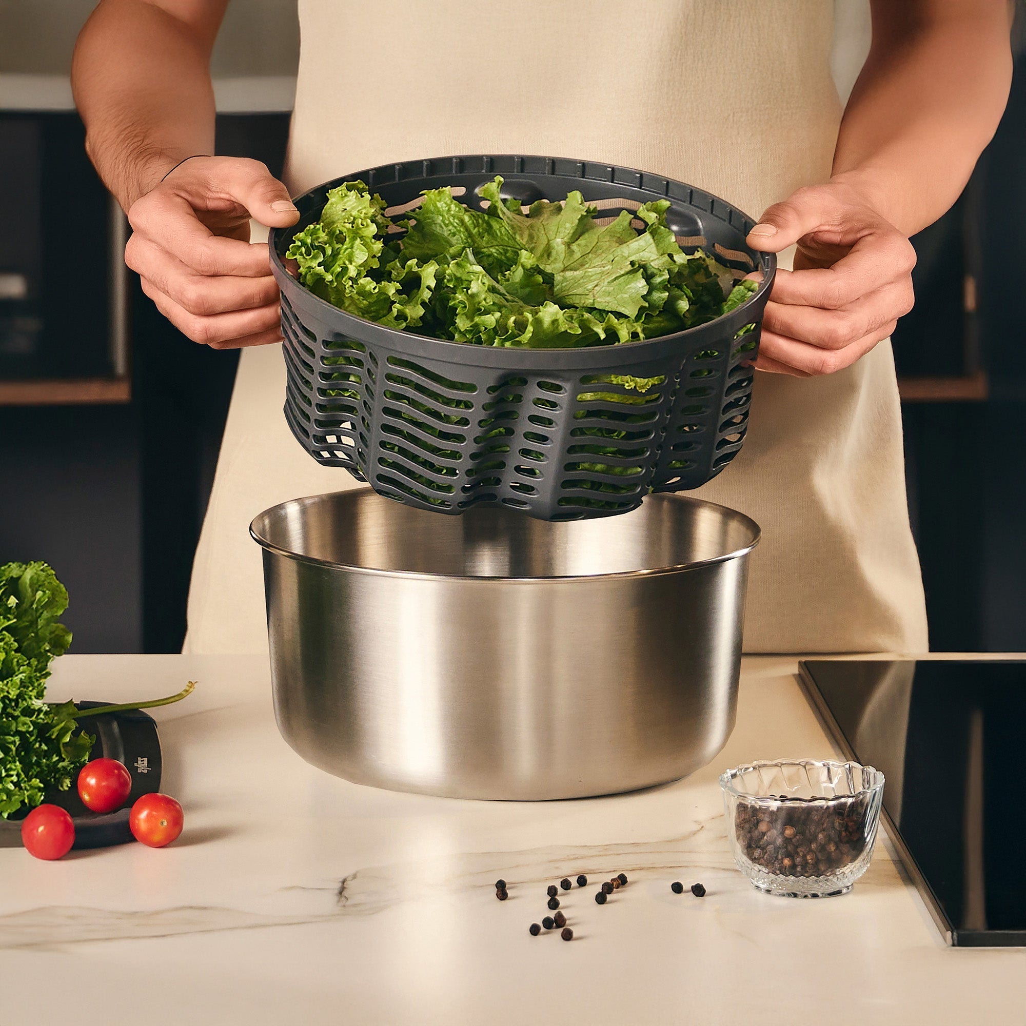A person using a salad spinner, lifting the perforated basket filled with leafy greens over a stainless steel bowl on a kitchen counter.