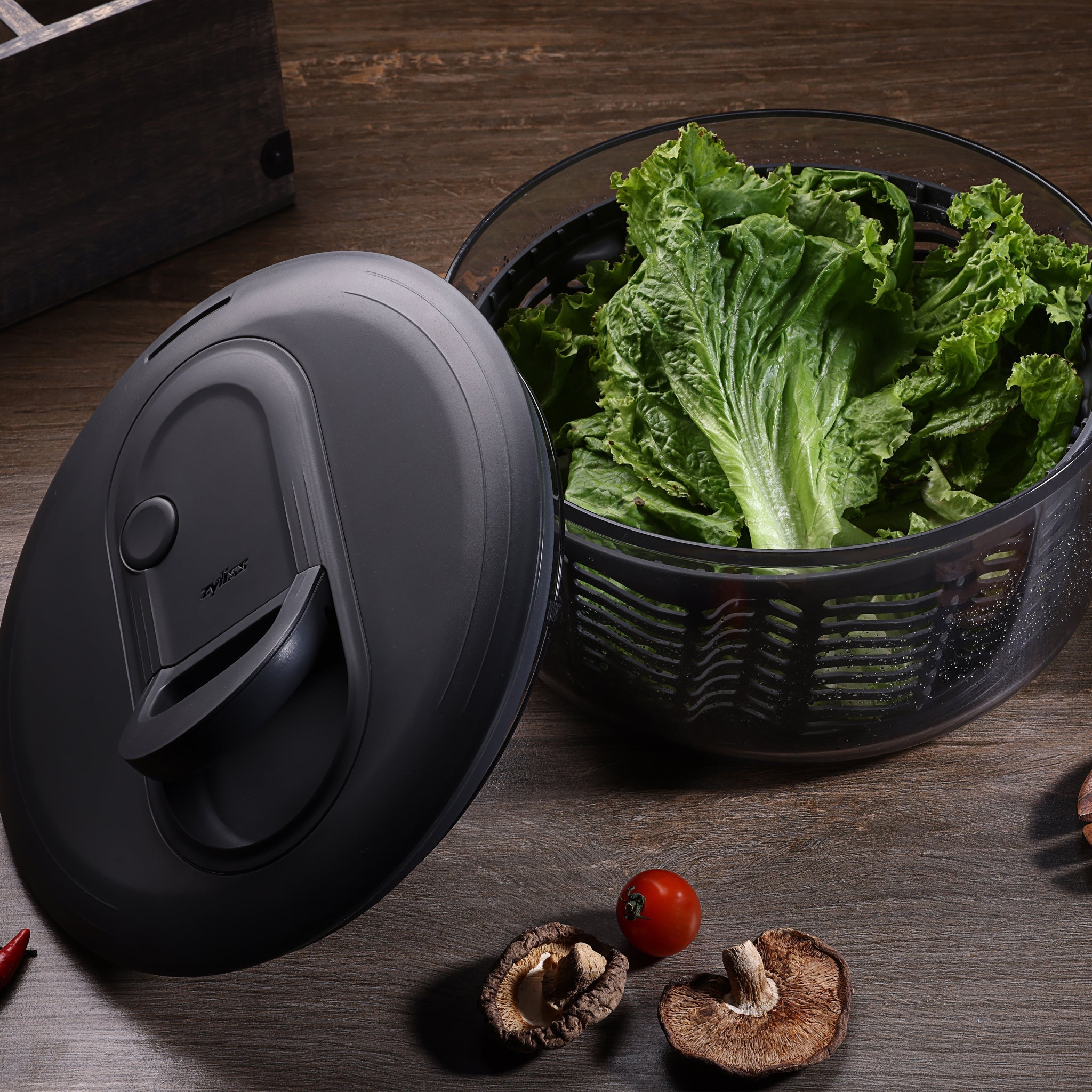 A fresh salad with greens, radishes, cucumbers, and carrots in a glass bowl, a white plate with salad, a small bowl of seeds, and a green salad spinner on a marble surface.
