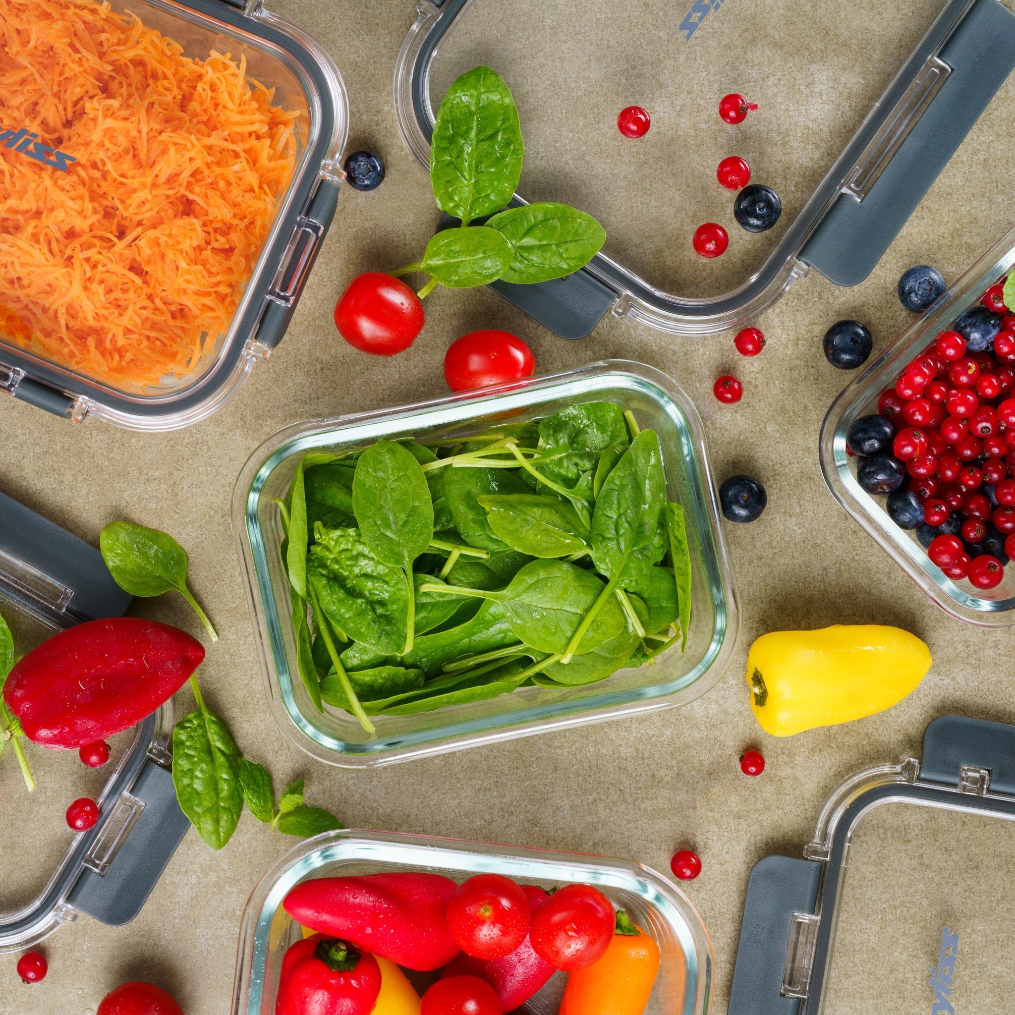 Glass containers filled with shredded carrots, spinach, and assorted berries, surrounded by cherry tomatoes and colorful mini peppers on a gray surface.