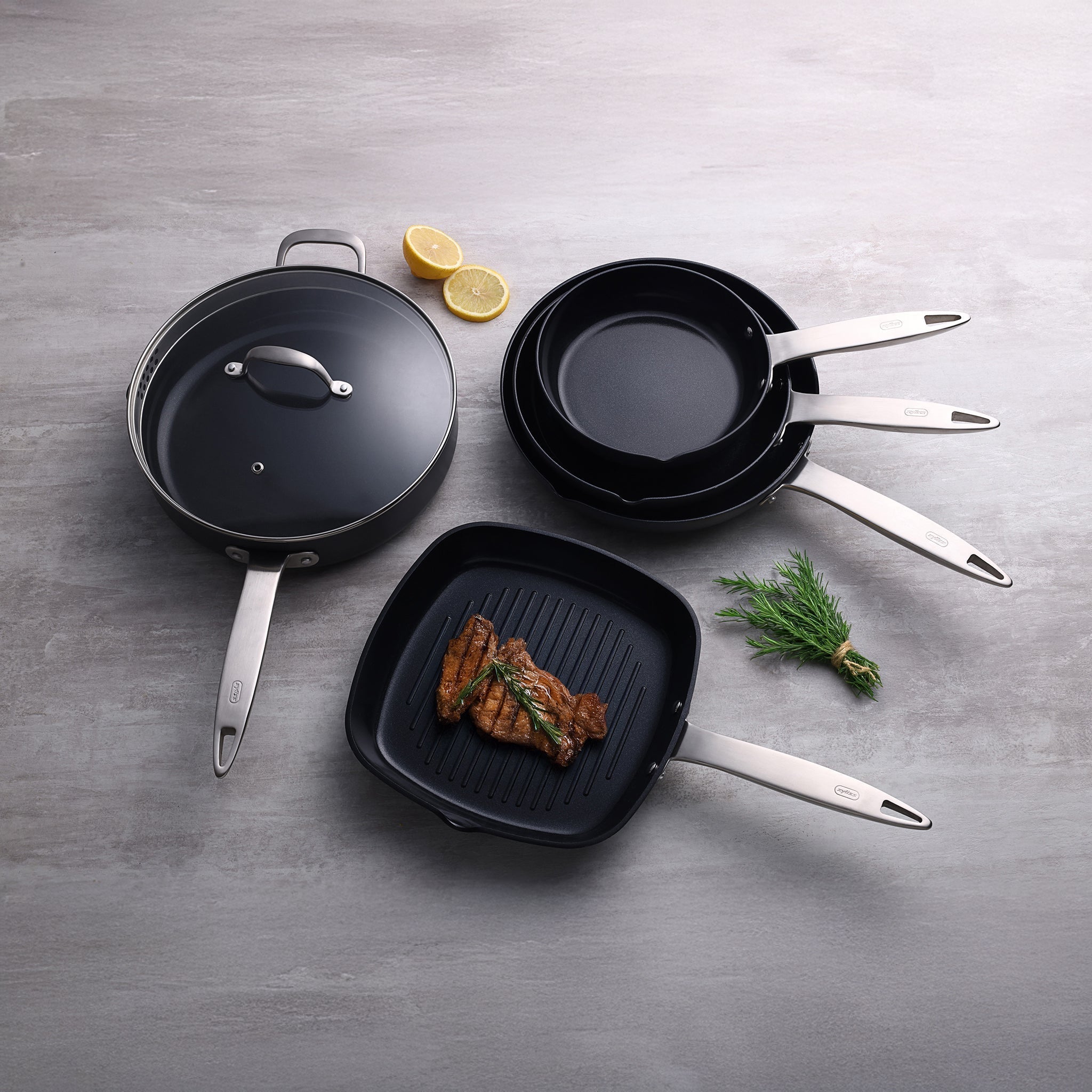 A set of non-stick frying pans and a grill pan with a lid, arranged on a gray surface. Grilled meat is in the grill pan, garnished with herbs. Two lemon halves and a rosemary sprig are beside the pans.