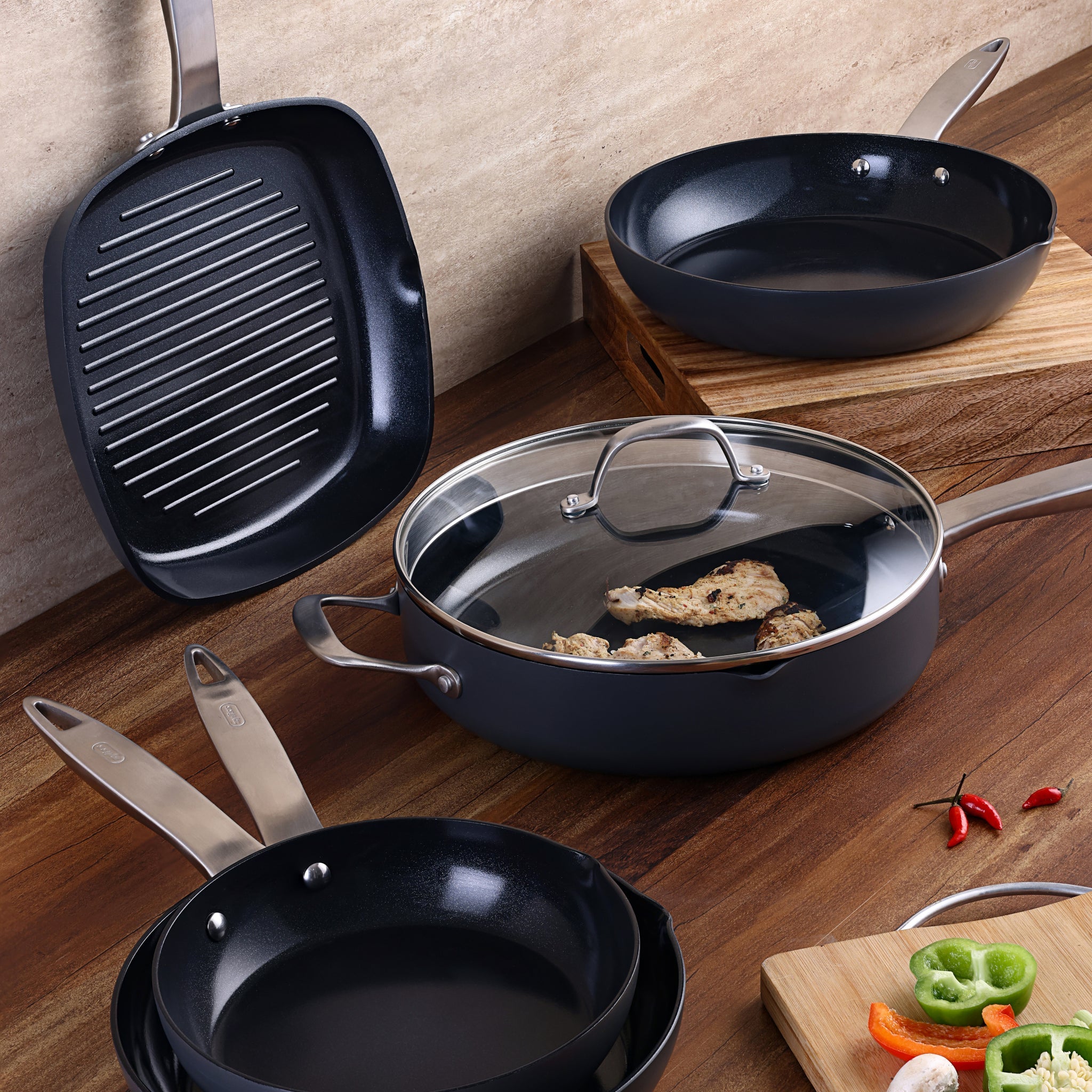 A set of non-stick cookware on a wooden countertop includes a grill pan, sauté pan with chicken and a glass lid, frying pans, and a cutting board with sliced bell peppers and chili peppers.