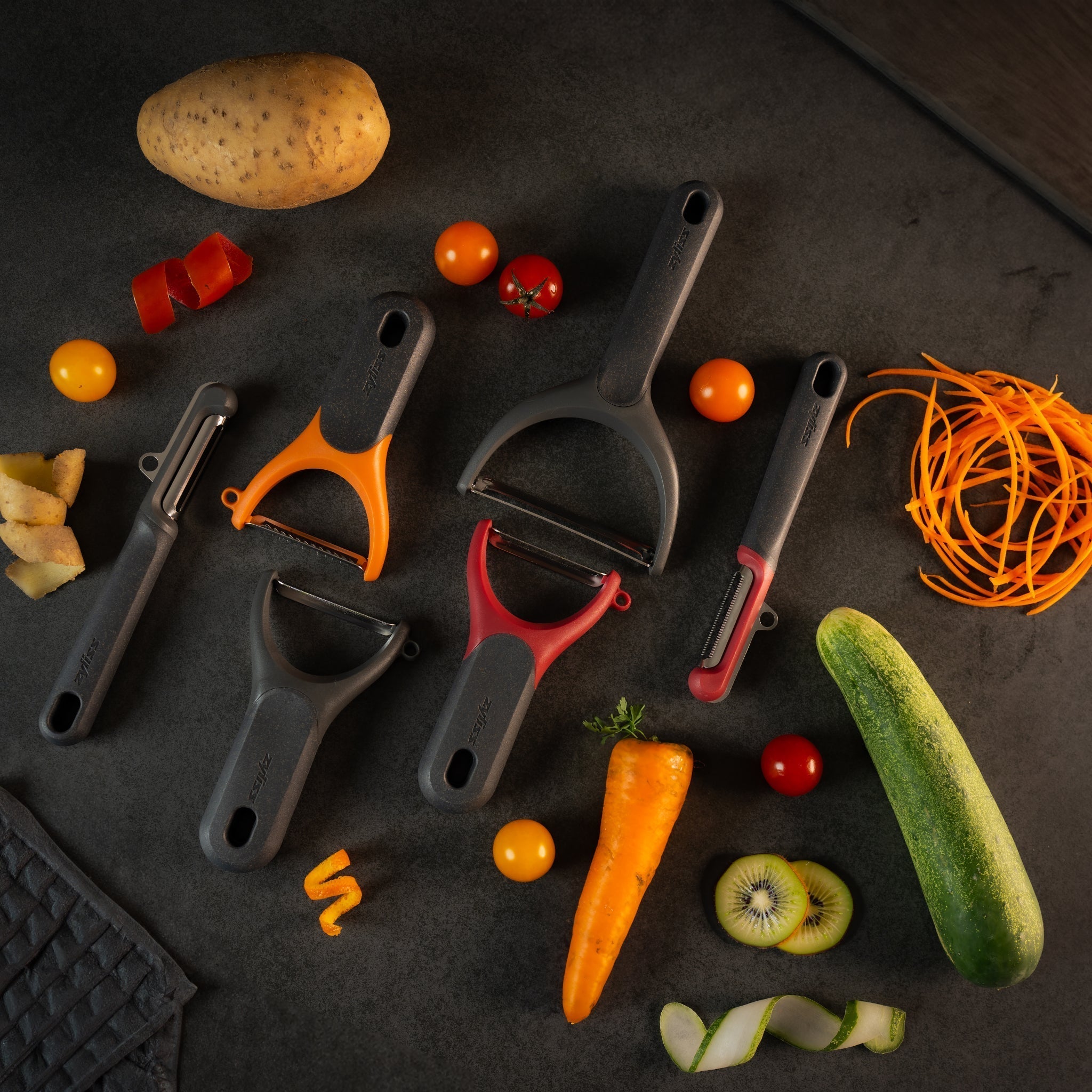 A variety of peelers are arranged on a dark surface with peeled and whole vegetables and fruits, including a potato, tomatoes, carrot, cucumber, kiwi, and orange zest.