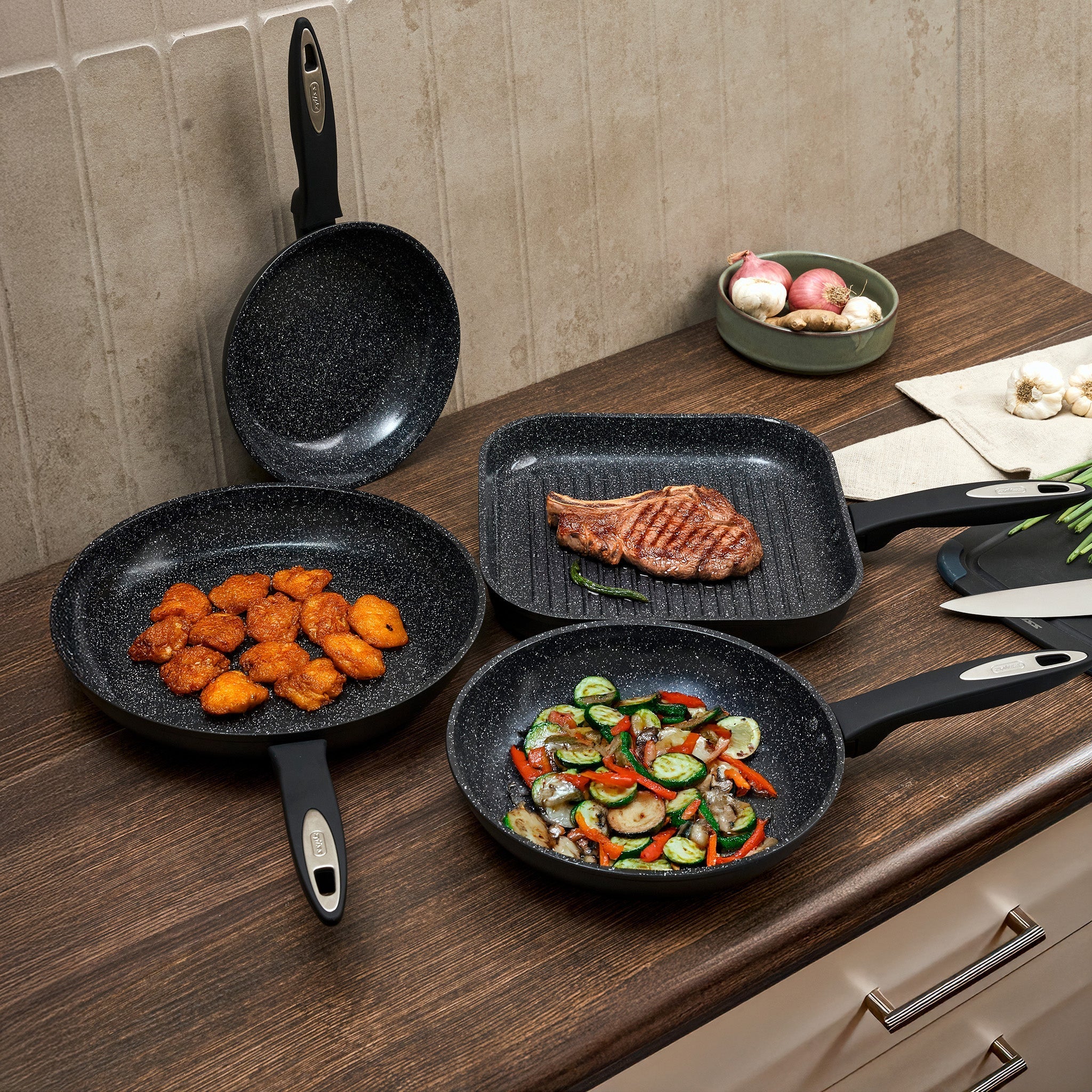 Three non-stick frying pans are displayed; one contains sautéed mixed vegetables, while the other two are stacked. Sliced potatoes are on a cutting board nearby, all set on a wooden surface.