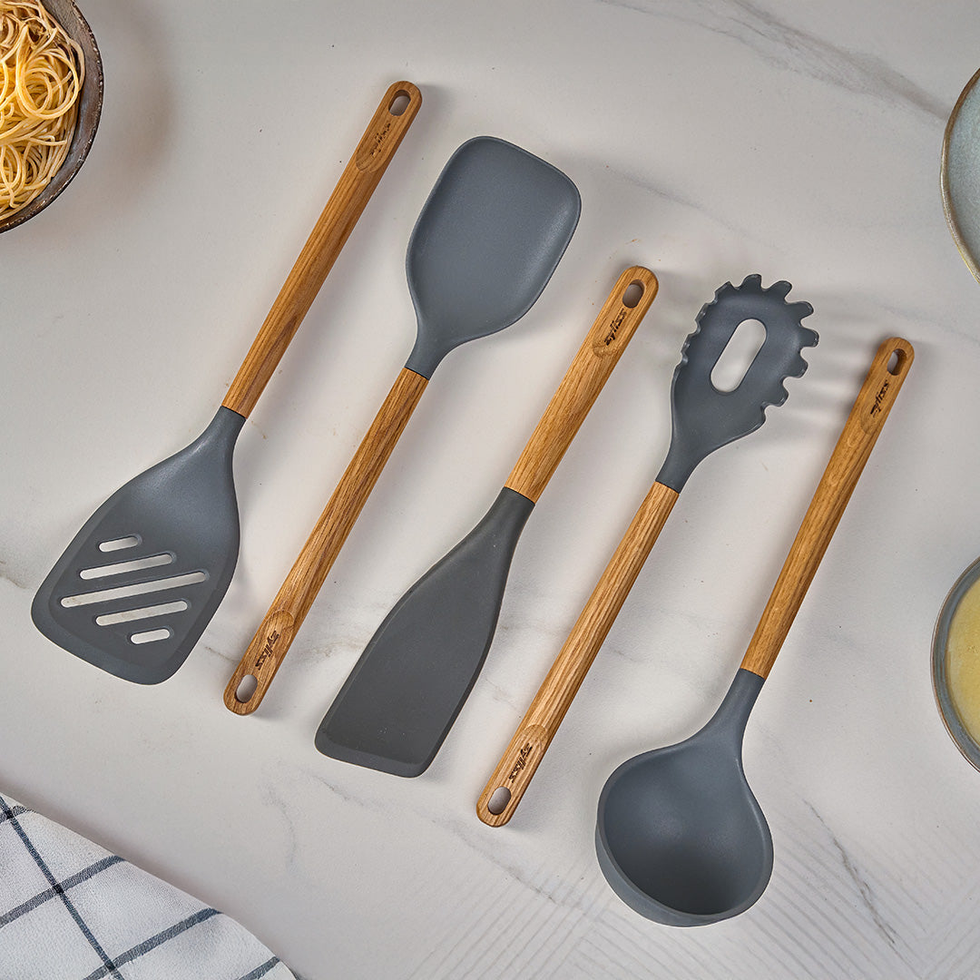 Four kitchen utensils with wooden handles and gray silicone heads, including a slotted spatula, regular spatula, pasta server, and ladle, are arranged neatly on a white marble surface.