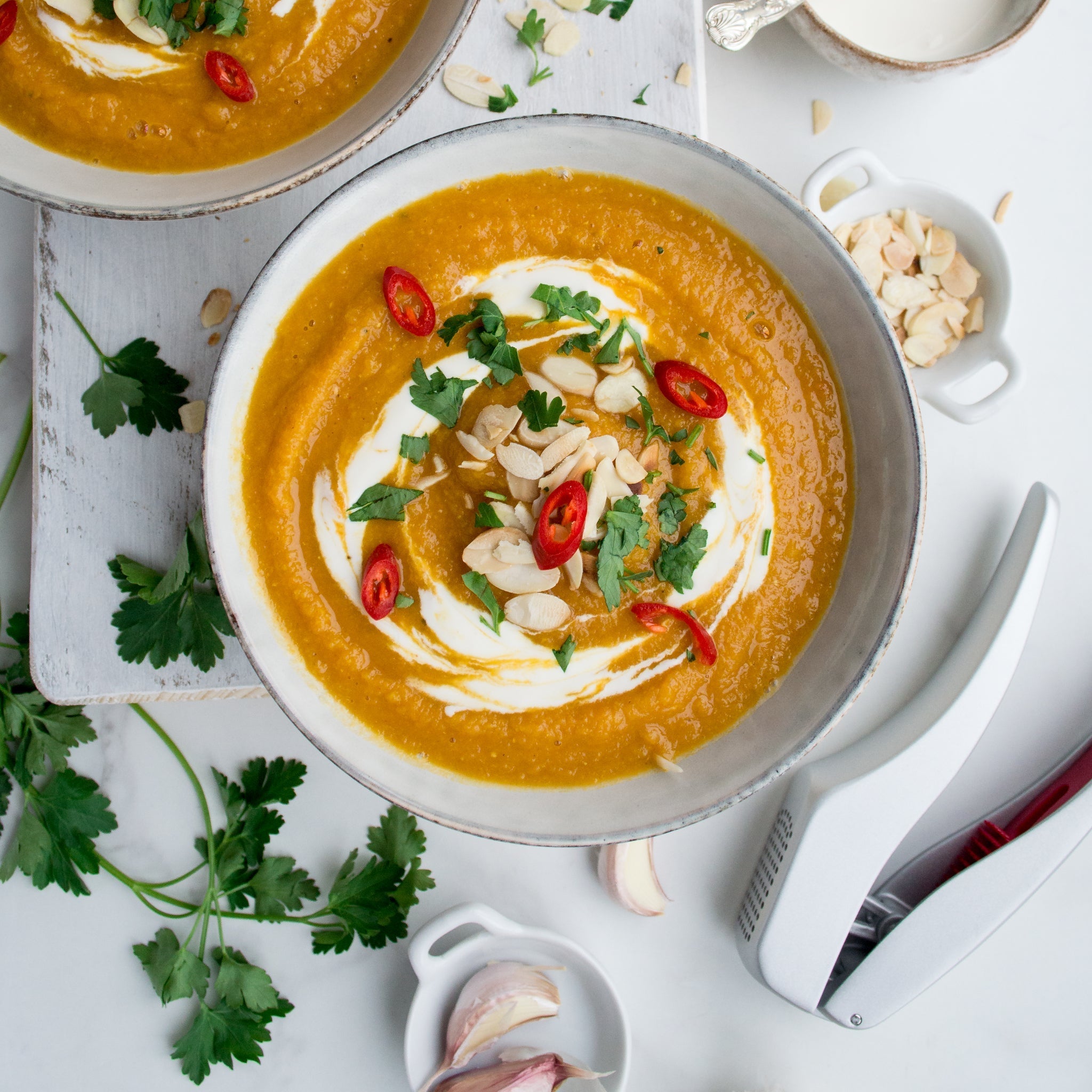 A bowl of creamy orange soup garnished with a swirl of cream, sliced red chili, fresh cilantro, and almond flakes, surrounded by parsley, garlic, and kitchen utensils on a white surface.