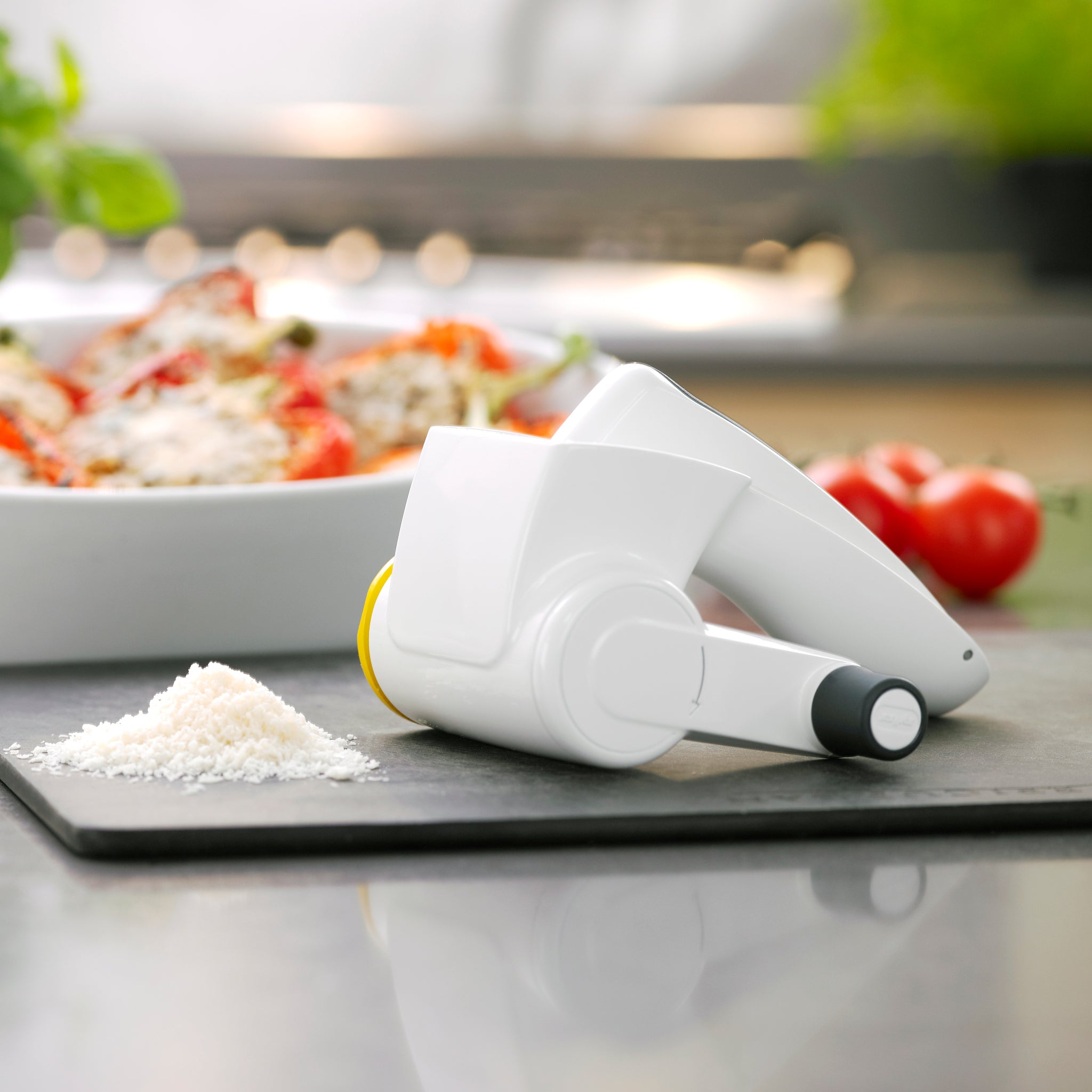 A white rotary cheese grater rests on a black surface near a pile of grated cheese, with a dish of food and fresh tomatoes blurred in the background.