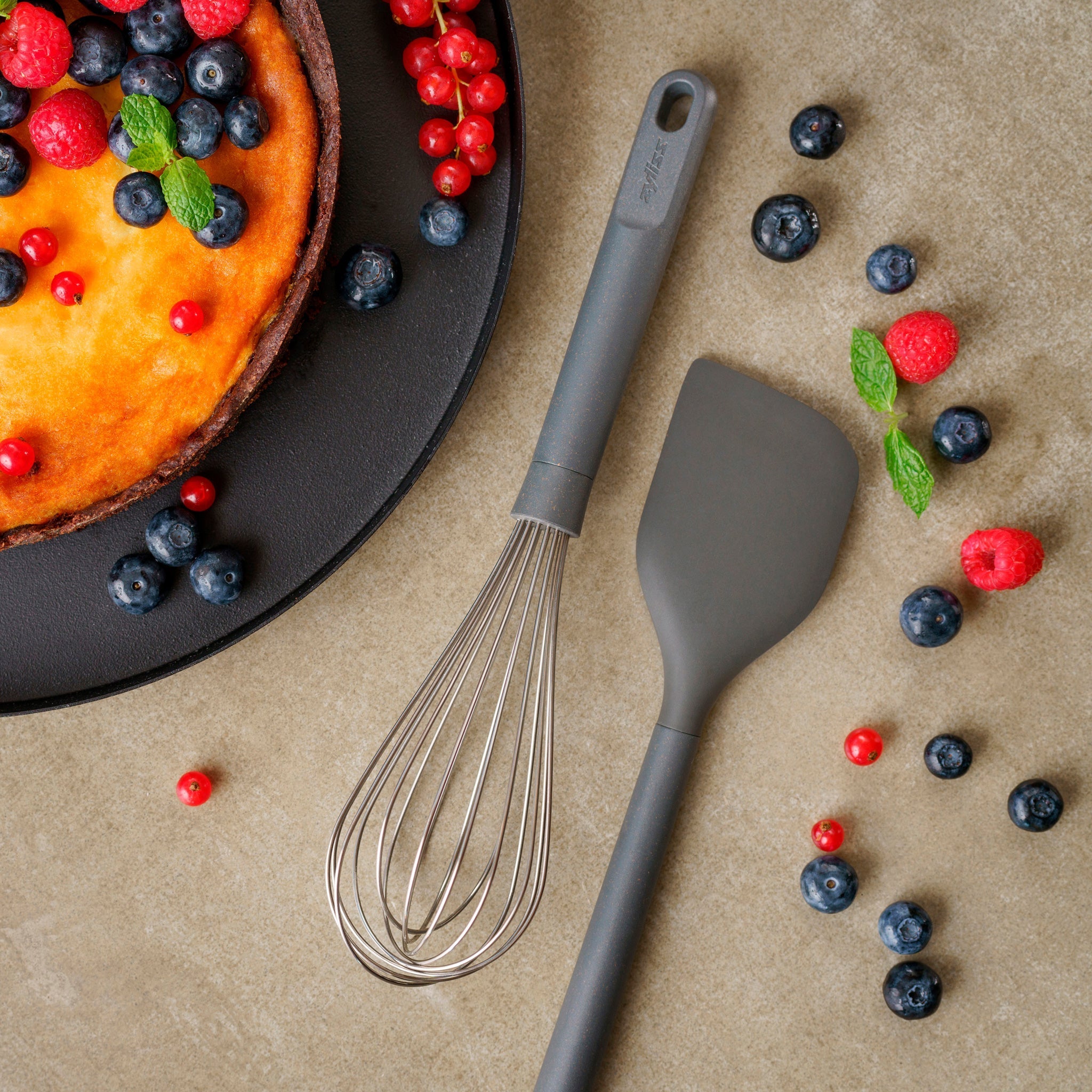 Cheesecake topped with blueberries, red currants, and mint leaves on a black plate. Nearby, a whisk and spatula are placed on a beige surface with scattered berries.