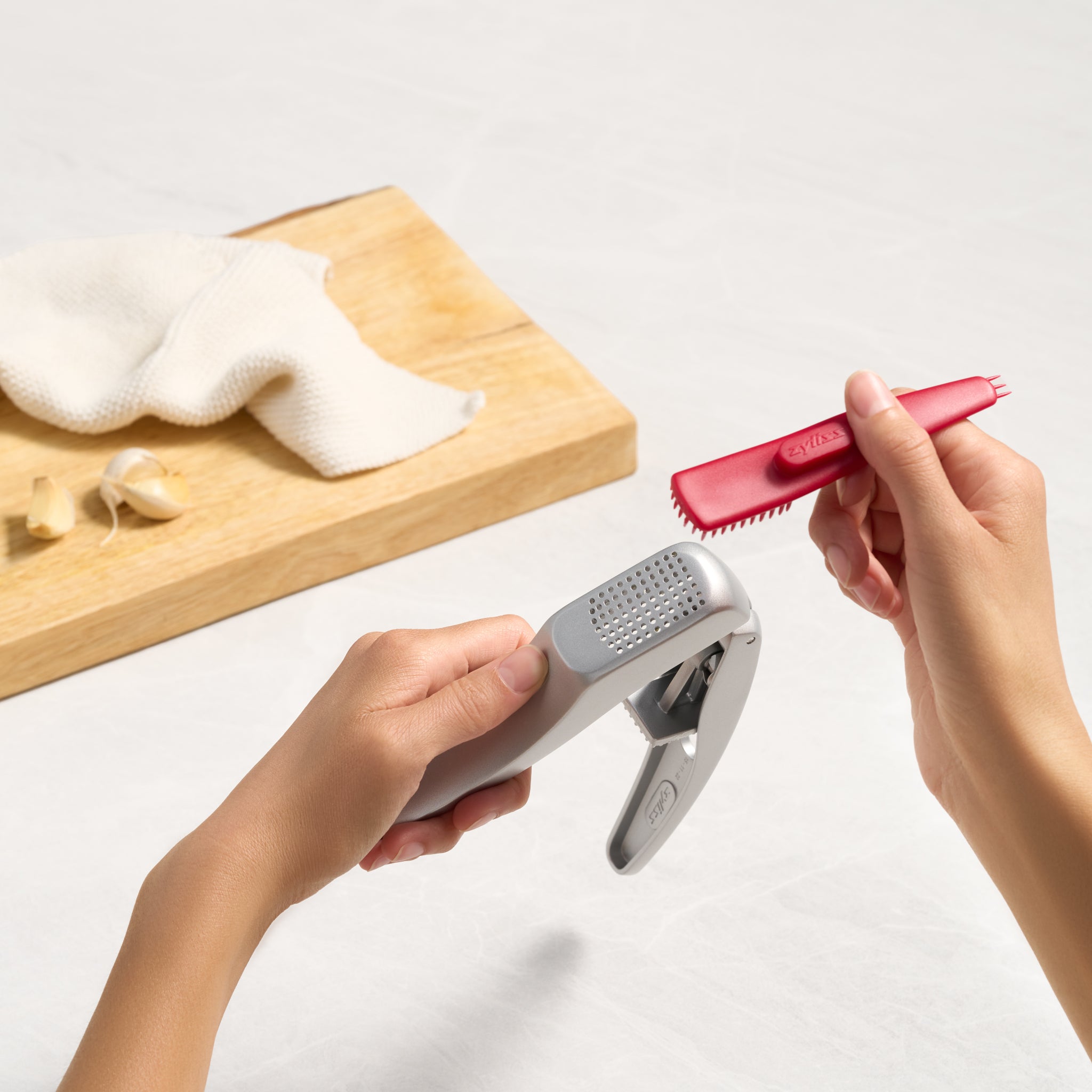 A person holds a garlic press in one hand and a red cleaning tool in the other, with garlic cloves, a cloth, and a wooden cutting board in the background.