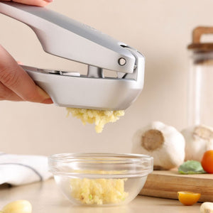 A hand uses the Zyliss Susi 4 Garlic Press by Zyliss, squeezing fresh garlic into a glass bowl on the kitchen counter, with garlic bulbs, a yellow tomato, and other kitchen items in the background.