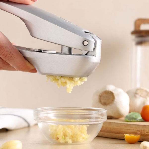 A hand uses the Zyliss Susi 4 Garlic Press by Zyliss, squeezing fresh garlic into a glass bowl on the kitchen counter, with garlic bulbs, a yellow tomato, and other kitchen items in the background.