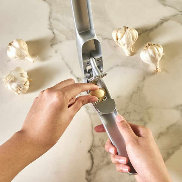 Someone places a garlic clove into the Zyliss Susi 4 Garlic Press, featuring ergonomic handles, on a marble countertop surrounded by whole garlic bulbs.