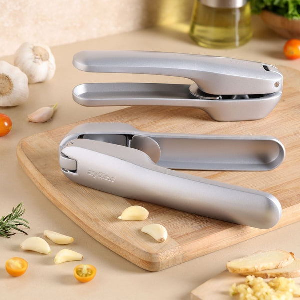 Two Zyliss Susi 4 Garlic Presses by Zyliss, featuring ergonomic handles, sit on a kitchen counter near chopped and whole garlic, cherry tomatoes, rosemary, oil, and a wooden cutting board holding peeled cloves and crushed garlic.