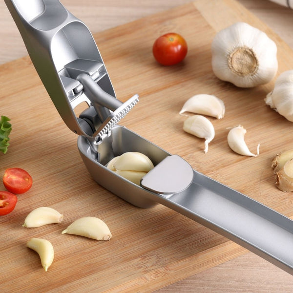 The Zyliss Susi 4 Garlic Press by Zyliss, filled with whole garlic cloves, sits on a wooden cutting board surrounded by tomatoes, garlic bulbs, loose cloves, and ginger. Its ergonomic handles ensure easy cleanup after use.
