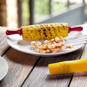 Grilled corn on the cob with Zyliss Interlocking Corn Holders rests on a white plate beside grilled shrimp skewers. A yellow corn-shaped butter spreader sits in the foreground on the wooden table. 