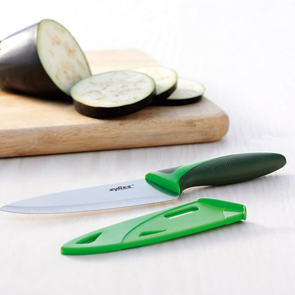 A green-handled knife from the Zyliss 3 Piece Knife Set with a matching sheath rests on a white surface beside a cutting board with sliced eggplant—an essential for any collection of versatile kitchen tools. 