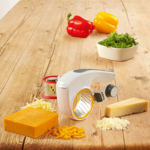 The Zyliss All Cheese Grater with interchangeable drums and sharp stainless steel blades rests on a wooden table, surrounded by cheddar and Parmesan blocks, shredded cheese, fresh greens, and colorful bell peppers in the background. 