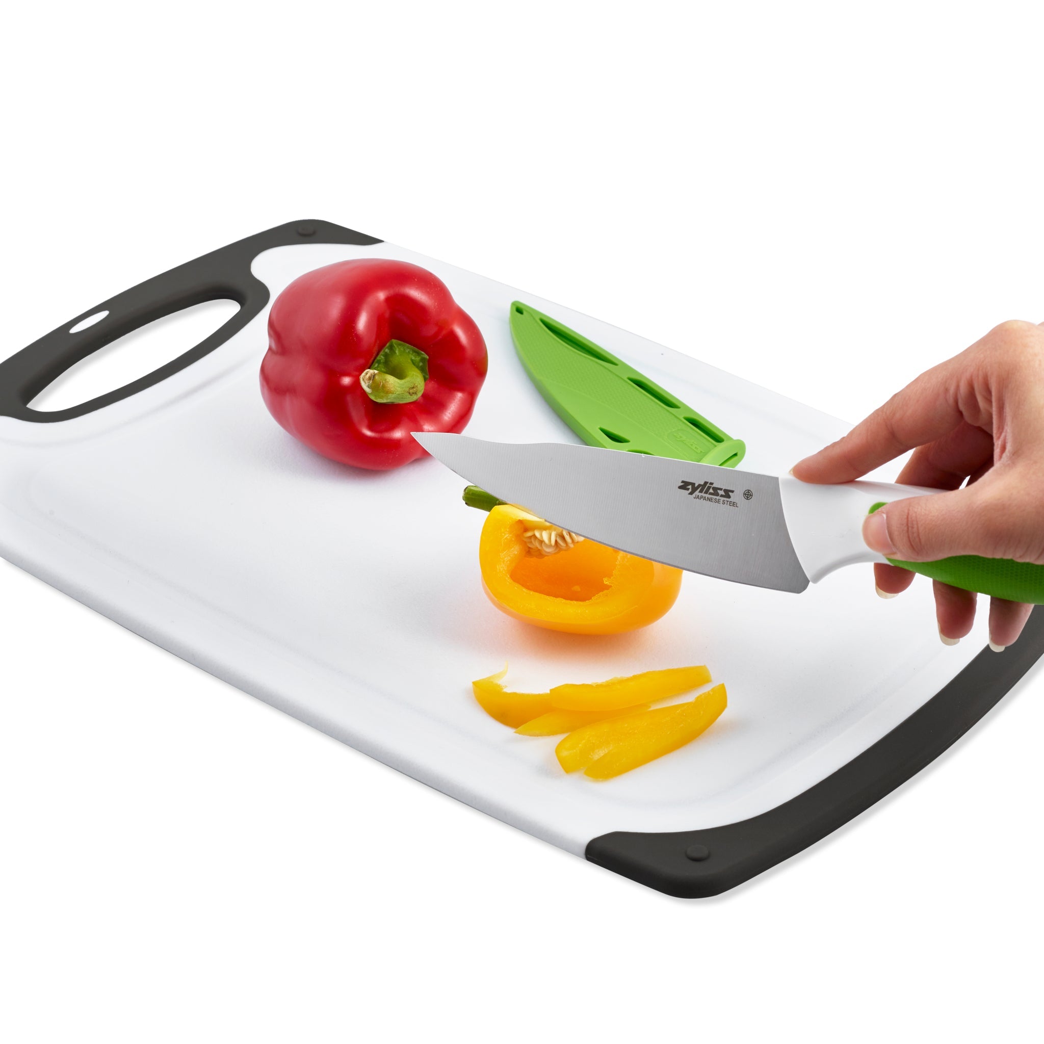 A hand slices a yellow bell pepper on the Zyliss 4 Piece Comfort Board and Knife Set, featuring a white board with black edge and a Japanese stainless steel blade for precision. Nearby are a red bell pepper, green knife sheath, and sliced yellow peppers.