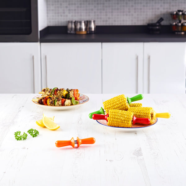 Grilled vegetable skewers and corn on the cob with Zyliss Interlocking Corn Holders sit on a white kitchen table, alongside lemon wedges, parsley, and a corn holder. A modern kitchen is visible in the background. 