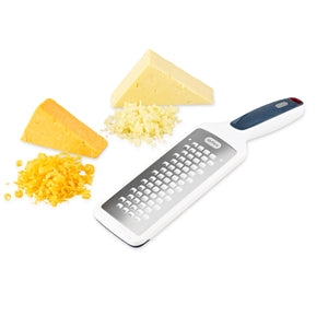 The Zyliss Smooth Glide Coarse Grater with a handle and stainless steel blade rests on a white surface beside two cheese wedges (yellow and white) and piles of freshly grated cheese. 