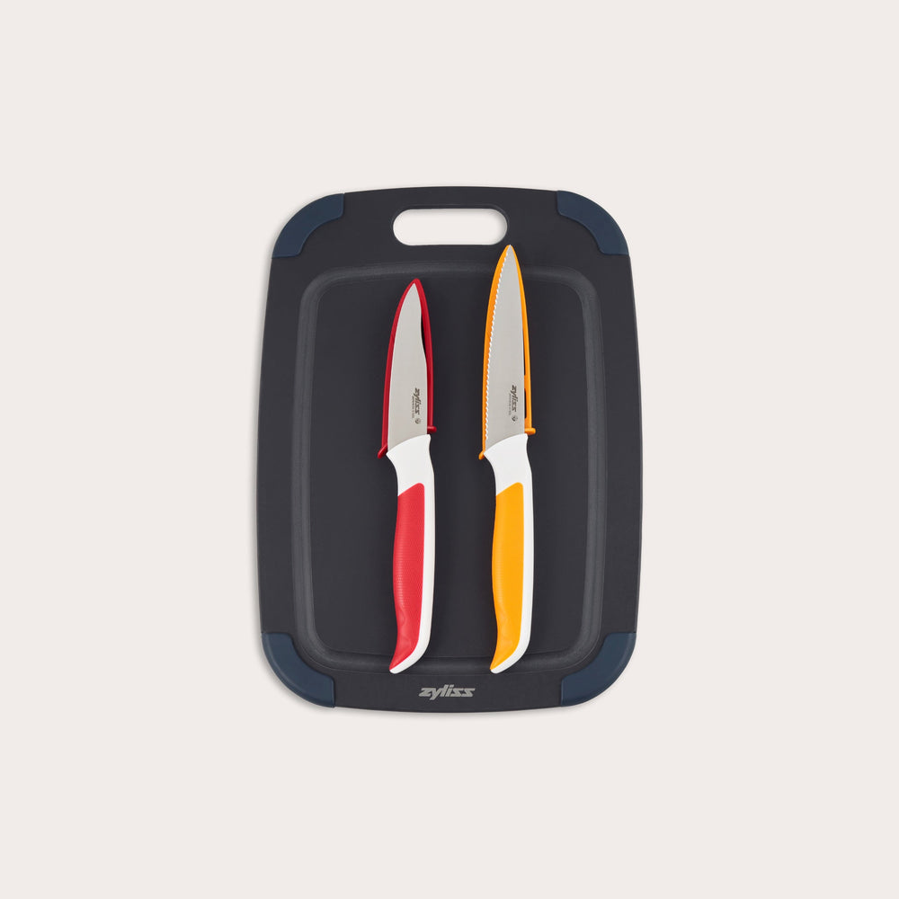 The Zyliss 3 Piece Wood Fibre Chopping Board & Knife Set includes a black cutting board with rounded corners and two knivesâ€”one red-handled and one orange-handledâ€”both with silver blades. 