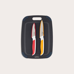 The Zyliss 3 Piece Wood Fibre Chopping Board & Knife Set includes a black cutting board with rounded corners and two knivesâ€”one red-handled and one orange-handledâ€”both with silver blades. 
