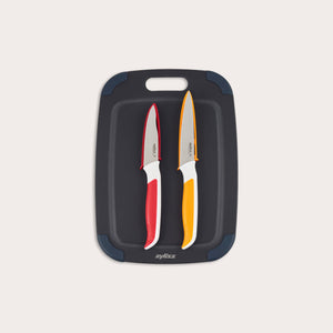 The Zyliss 3 Piece Wood Fibre Chopping Board & Knife Set includes a black cutting board with rounded corners and two knivesâ€”one red-handled and one orange-handledâ€”both with silver blades. 