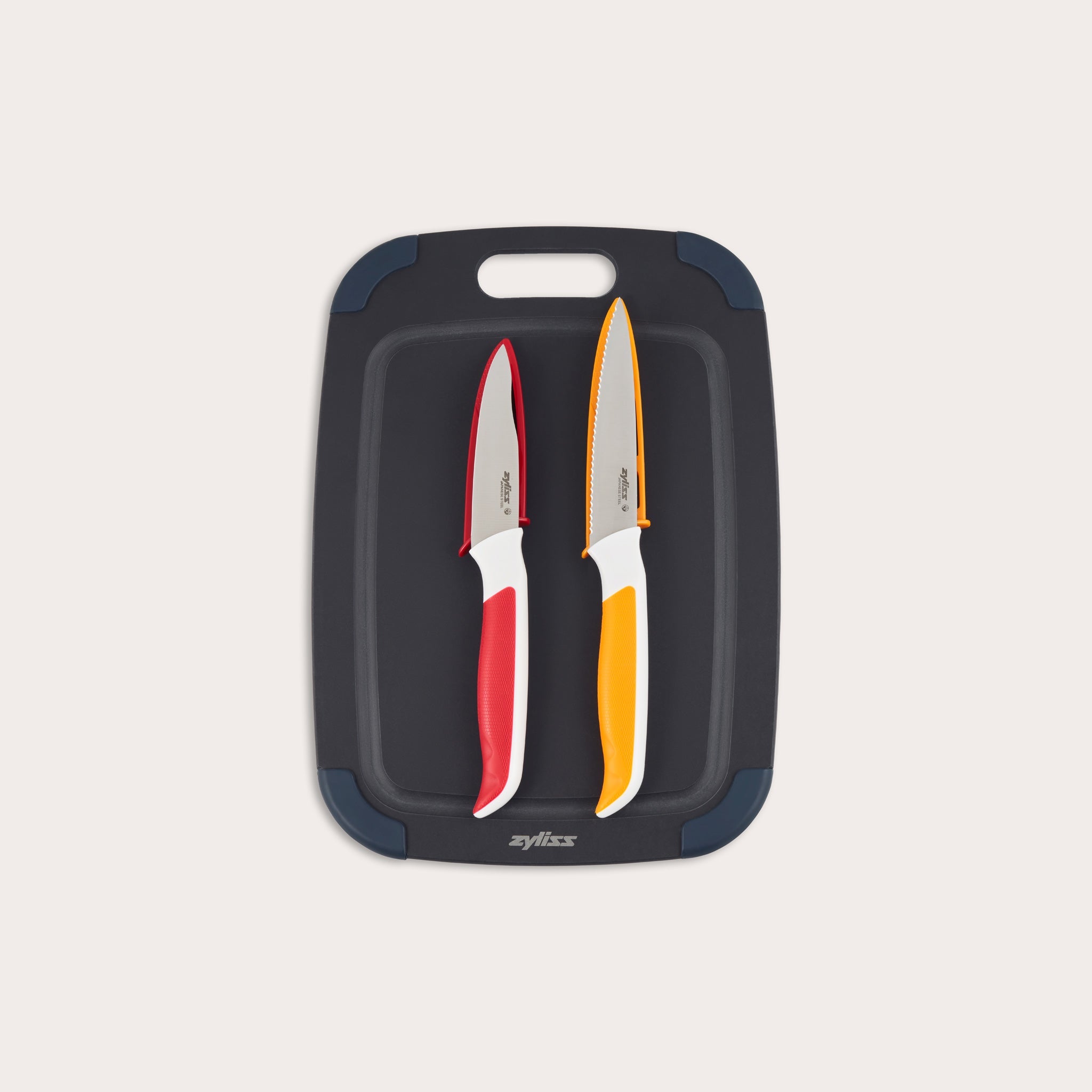 The Zyliss 3 Piece Wood Fibre Chopping Board & Knife Set includes a black cutting board with rounded corners and two knivesâ€”one red-handled and one orange-handledâ€”both with silver blades. 