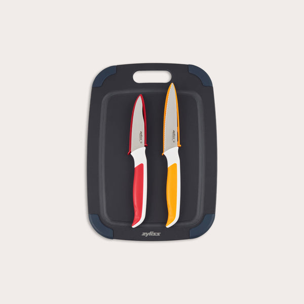 The Zyliss 3 Piece Wood Fibre Chopping Board & Knife Set includes a black cutting board with rounded corners and two knivesâ€”one red-handled and one orange-handledâ€”both with silver blades. 