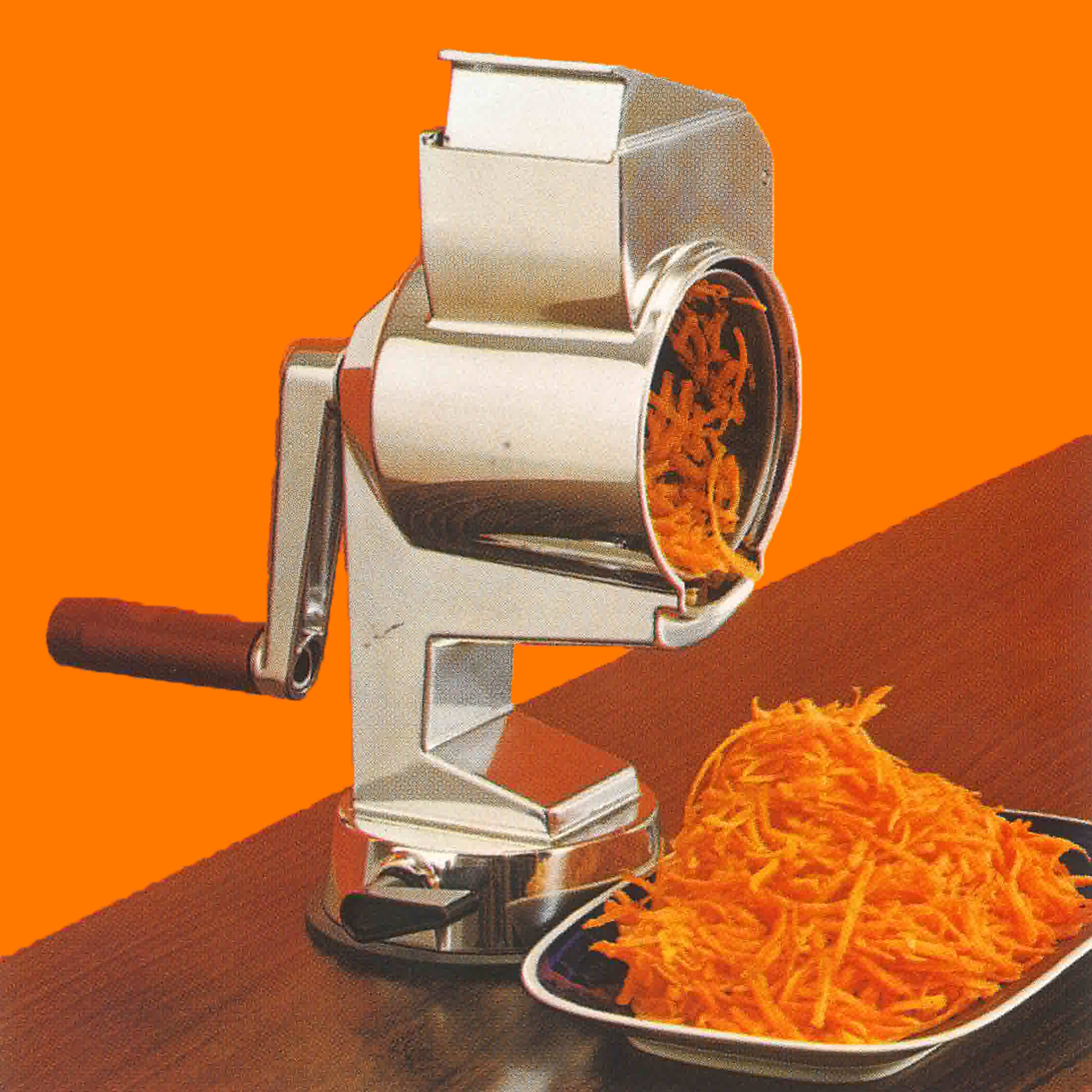 A metal rotary grater with a wooden handle is grating carrots onto a plate on a wooden table, set against a solid orange background.