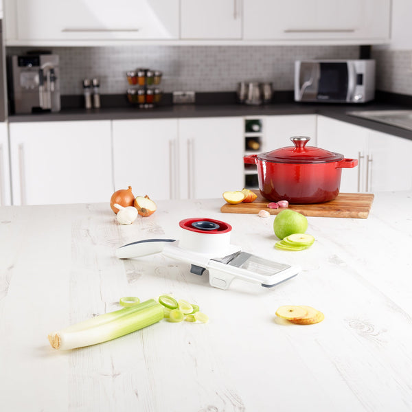 A white kitchen with vegetables and the Zyliss Easy Control Handheld Slicer on the counter. Sliced leek and apple rest by its food holder, while a red pot sits on a wooden board nearby, surrounded by more vegetables. 