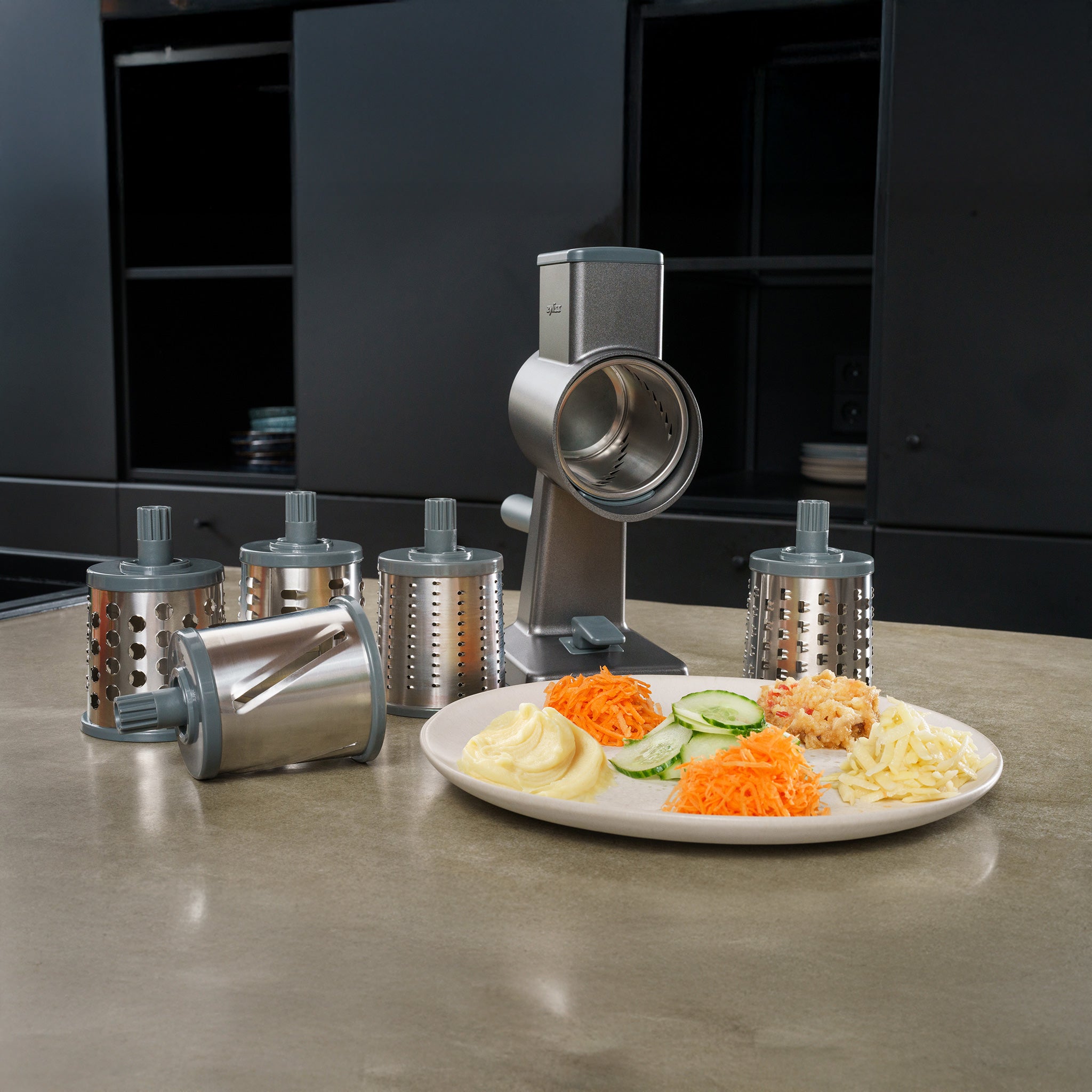 A rotary grater with multiple interchangeable drum blades is displayed on a kitchen counter, next to a plate with sliced cucumbers, shredded carrots, mashed potatoes, and grated cheese.