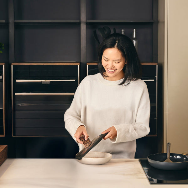 A woman in a white sweater smiles in a modern kitchen as she uses the Zyliss Rasp Grater to grate chocolate over a bowl on the counter. A frying pan rests on the stove, and dark shelves are visible in the background. 