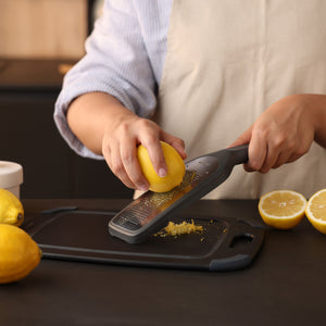A person wearing a beige apron uses the ultra-sharp Zyliss Fine Grater to zest a lemon over a black cutting board, surrounded by lemons on a kitchen counter—a true sustainable kitchen essential. 