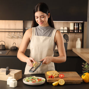 A woman in a beige apron uses the Zyliss Coarse Grater to grate cheese onto a salad in a modern kitchen. Fresh vegetables, a lemon, and a cheese block are on the cutting board as she smiles and focuses on her task. 