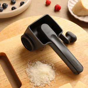 The Zyliss Professional Rotary Cheese Grater in black, featuring a stainless steel drum, sits on a wooden cutting board with grated cheese, cheese blocks, berries, and a wedge of cheese. Its dishwasher safe for easy cleaning.