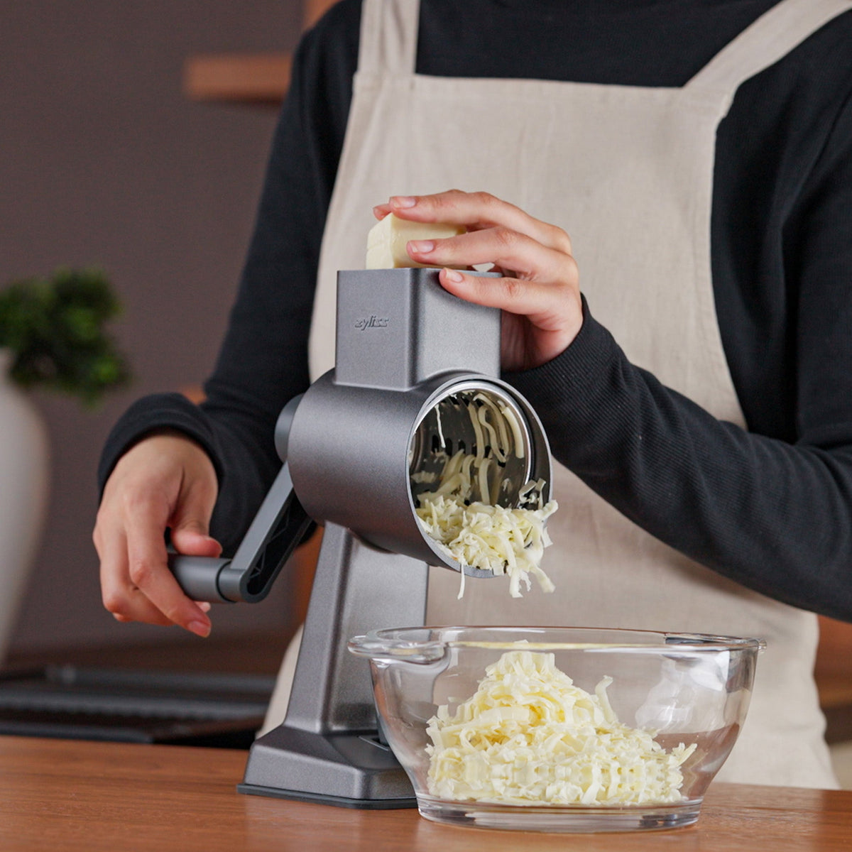 #drumgratersingle #râpeàtamboursimple A person in a beige apron uses the Zyliss Gourmet Drum Grater to shred cheese into a glass bowl on a wooden countertop.