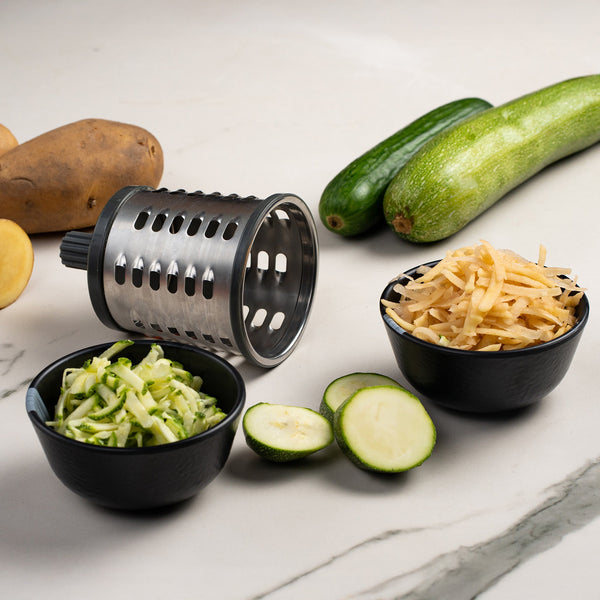 #drumgratersingle The Zyliss Gourmet Drum Grater is shown with shredded zucchini and potatoes in black bowls, alongside fresh zucchini slices, whole potatoes, and whole zucchini on a white marble surface.