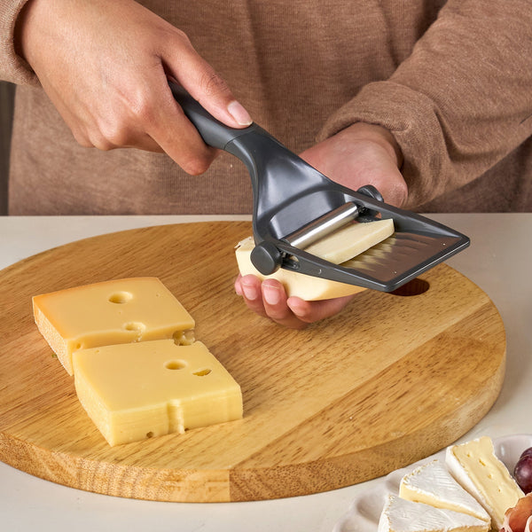 A person uses the Zyliss Dial & Slice Cheese Slicer, featuring an adjustable stainless steel blade, to cut cheese slices from a block on a round wooden cutting board while other cheese pieces are nearby.