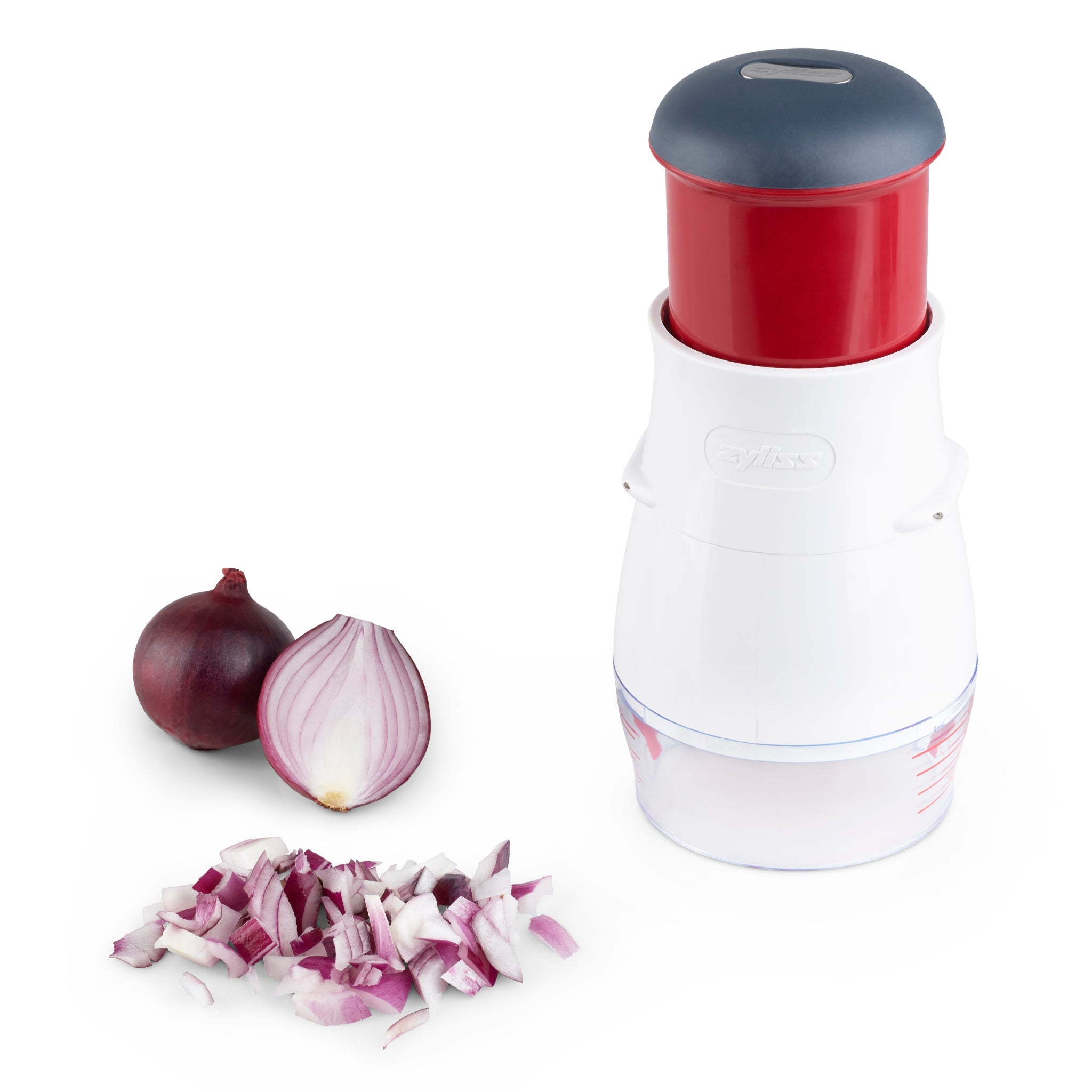 A manual onion chopper with a red and gray top, white body, and clear base is shown next to a whole red onion, a halved onion, and chopped onion pieces on a white surface.
