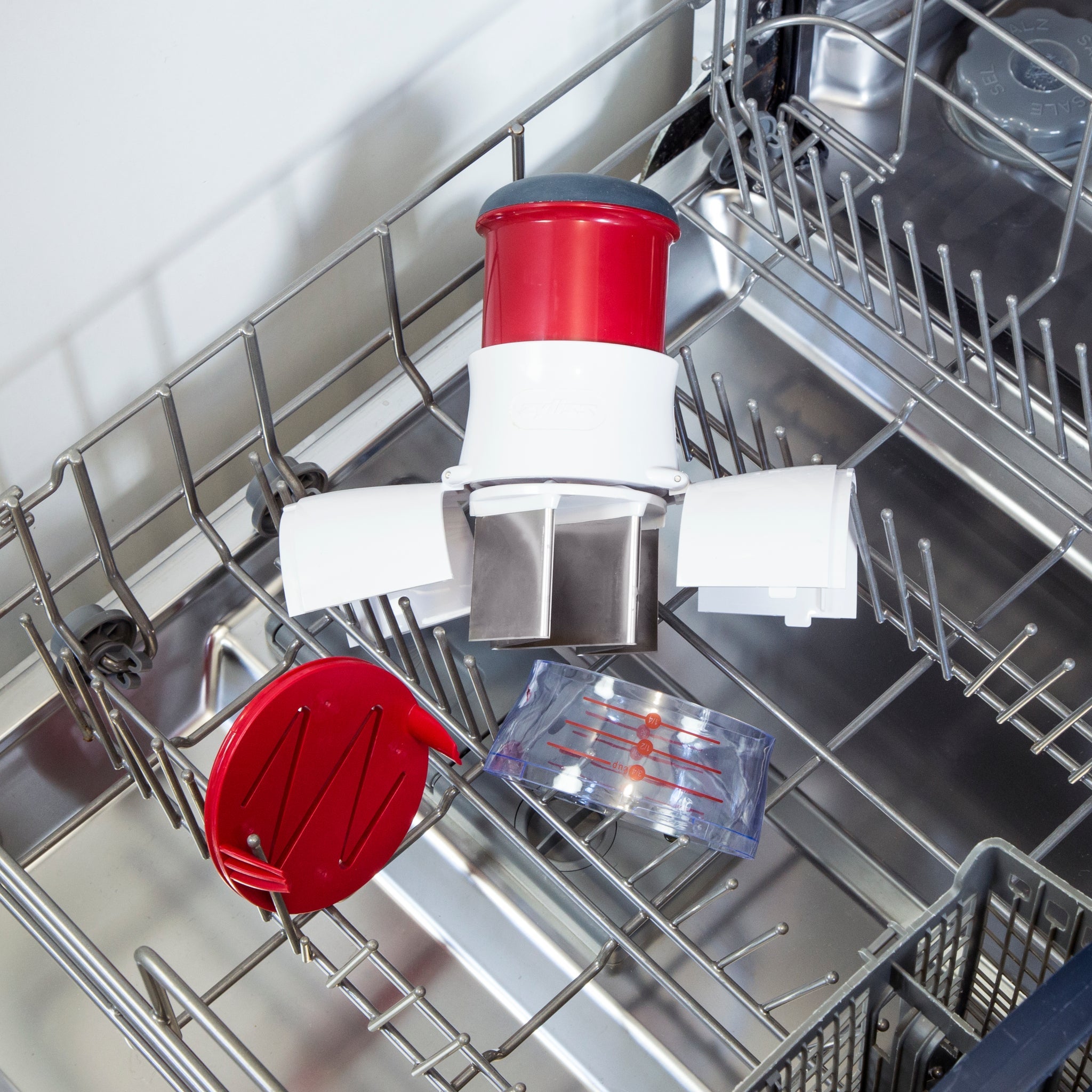 A red and white kitchen gadget with metal blades and a clear container is placed on the top rack of an open dishwasher, next to a matching red lid.
