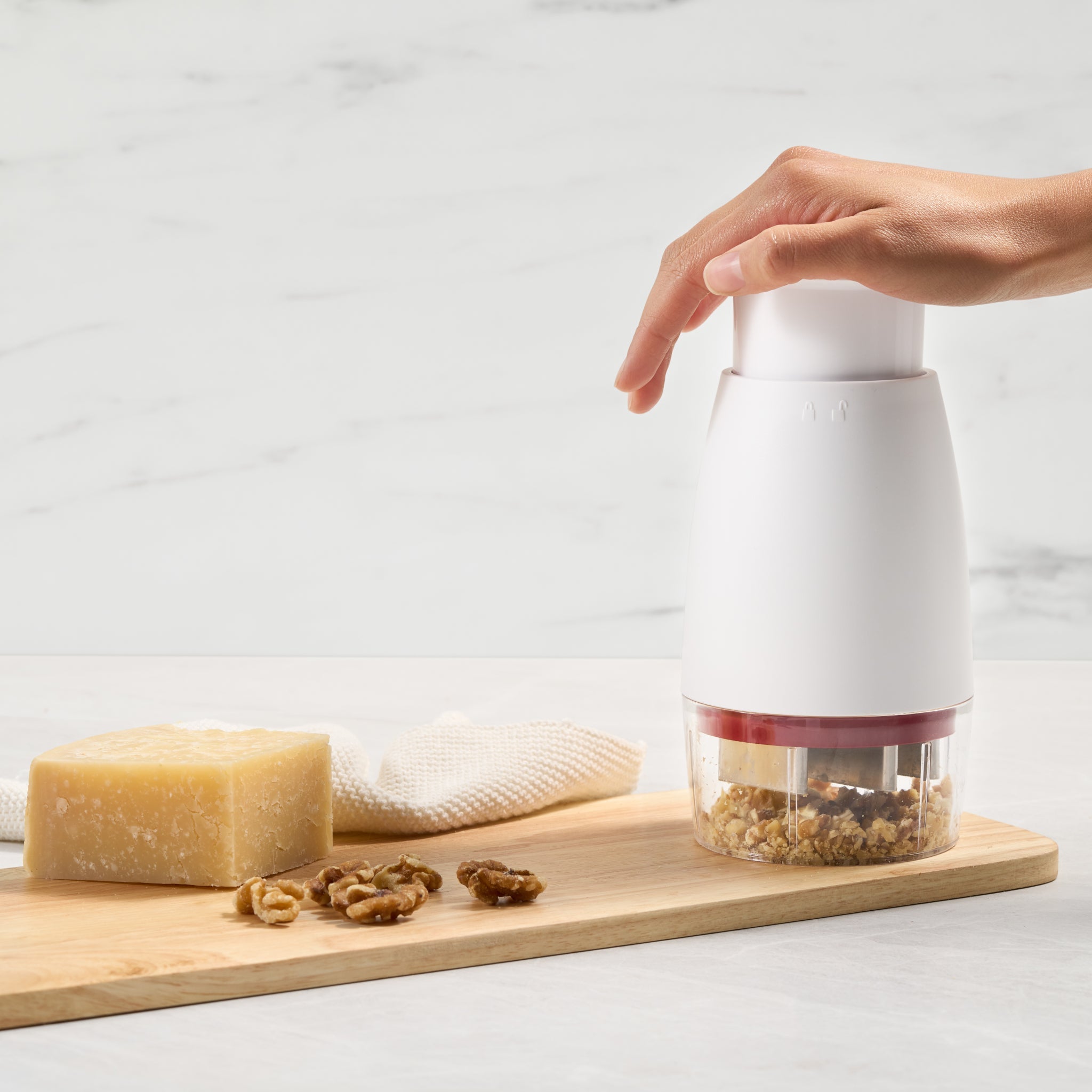 A hand presses down on a white food chopper, chopping walnuts on a wooden board. A block of cheese, some loose walnuts, and a cloth are also on the board, all set against a light background.