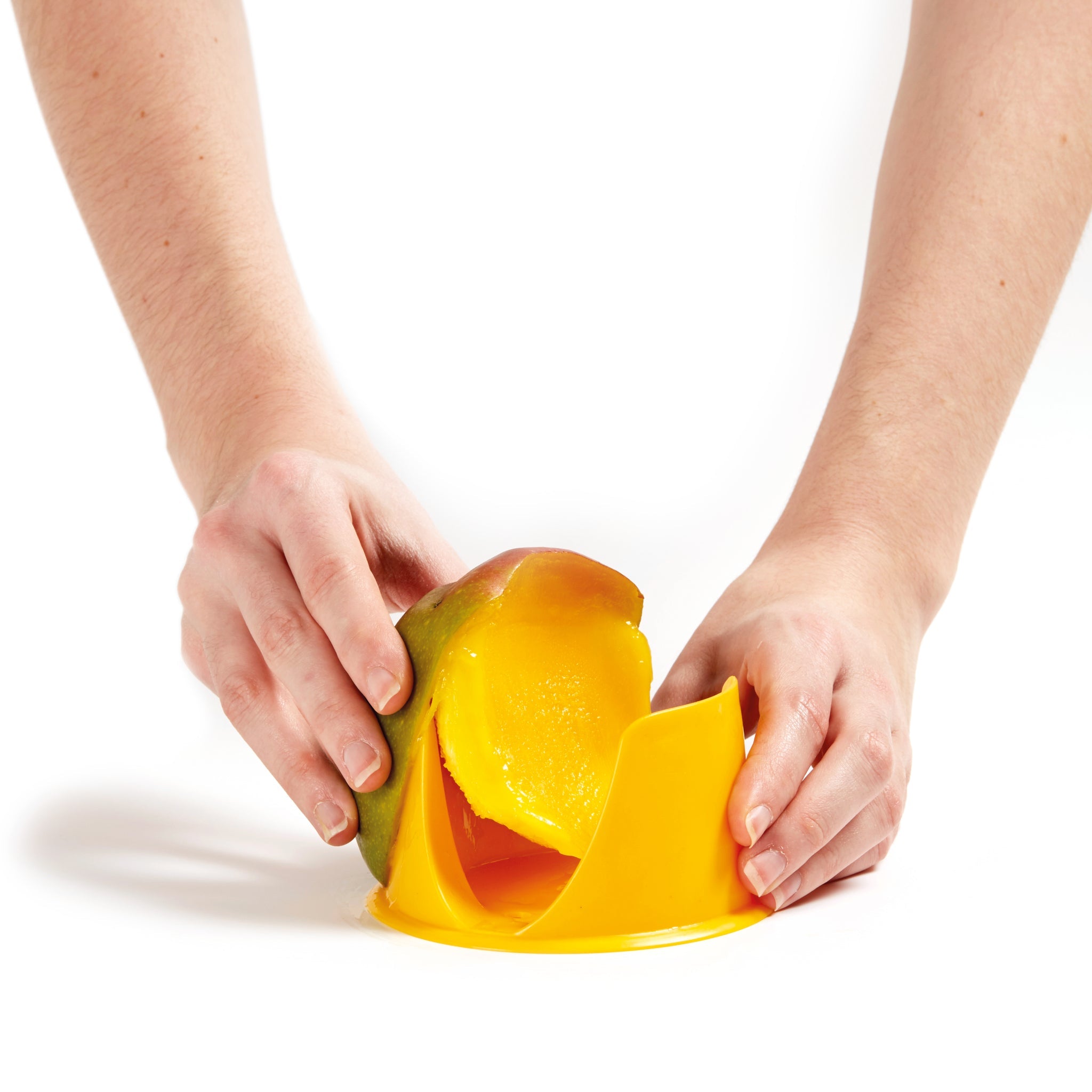 A person uses a yellow plastic tool to slice a mango, holding the fruit steady with one hand while pushing it through the slicer with the other, against a white background.