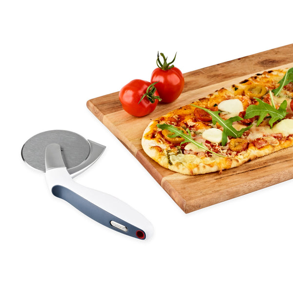 A rectangular arugula and cheese pizza sits on a wooden board beside two tomatoes, with the Zyliss Sharp Edge Pizza Cutter Wheel, featuring an ergonomic handle and stainless steel blade, nearby on a white background. 
