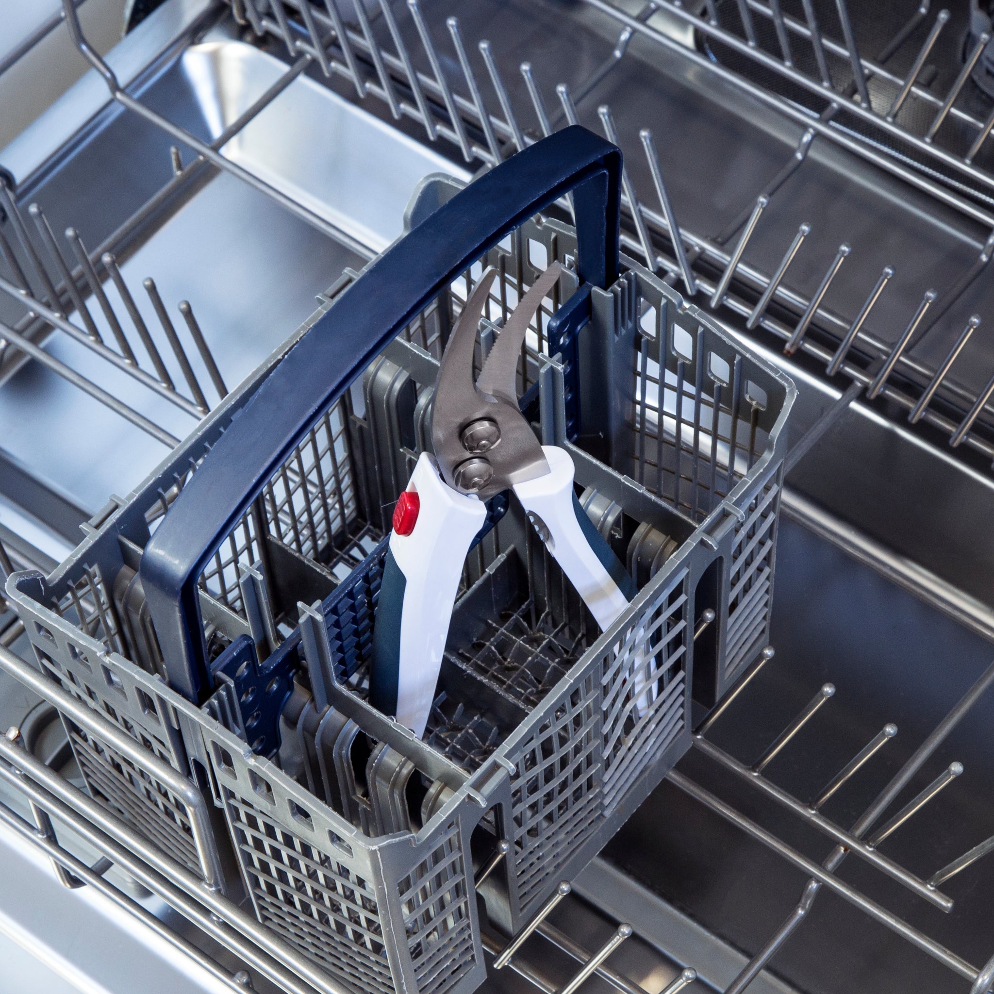 A pair of garden pruning shears with white handles placed inside the utensil basket of an open, empty dishwasher.